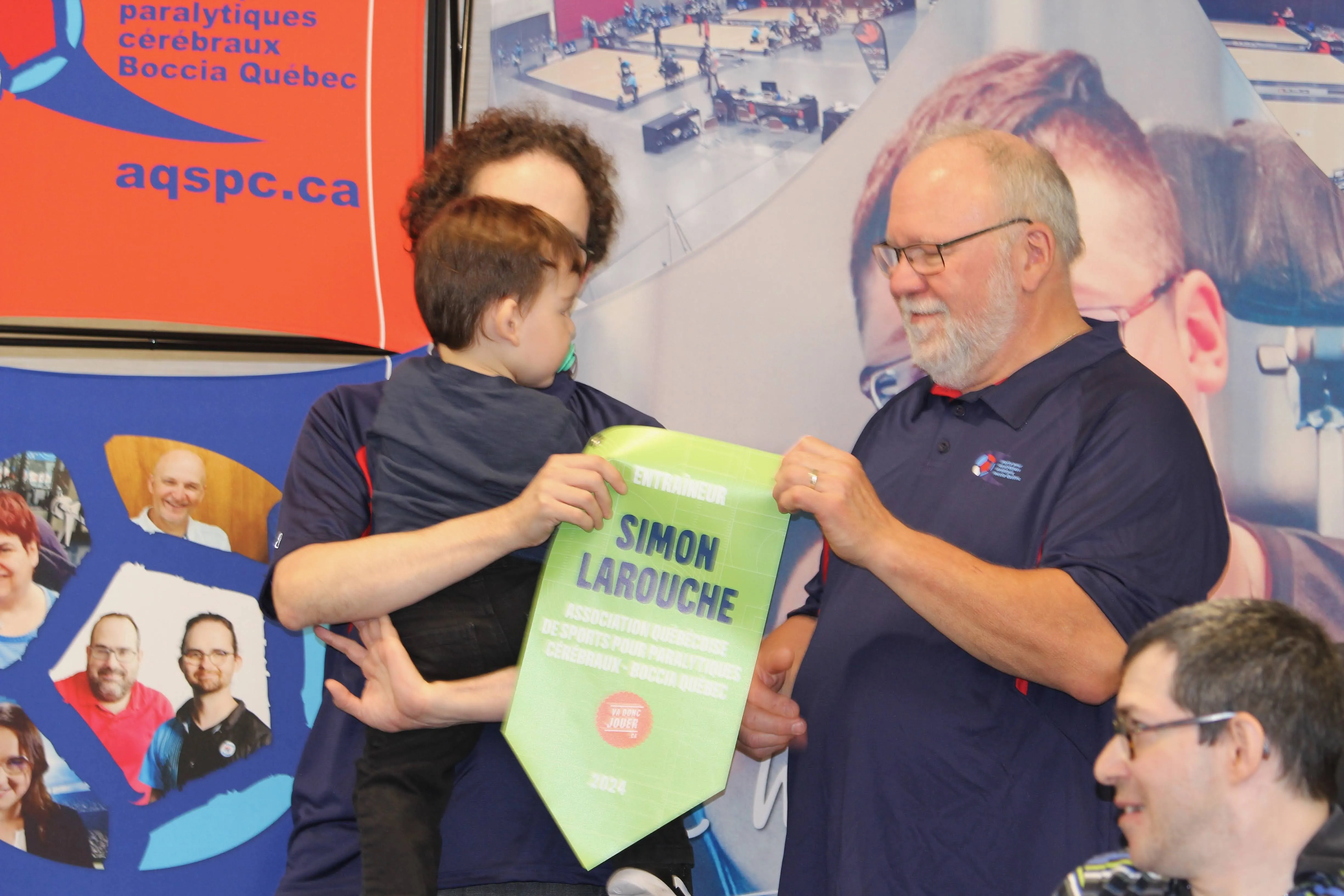 Simon, tenant son fils dans ses bras, reçoit son drapeau de meilleur entraîneur des mains de François Bureau, président de l'AQSPC.